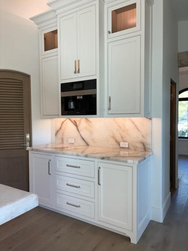 White kitchen cabinets with marble backsplash, built-in coffee maker, and wooden floor.