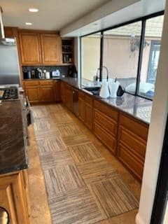 Kitchen with wood cabinets, dark countertops, and a long, patterned rug. Large window and outdoor view.