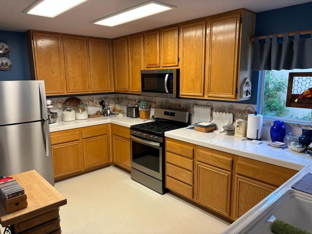 A well-lit kitchen with light oak cabinets, stainless steel appliances, and white countertops.