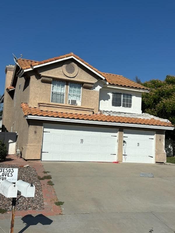 Two-story tan house with white garage doors and red tile roof, driveway, and mailbox in front.