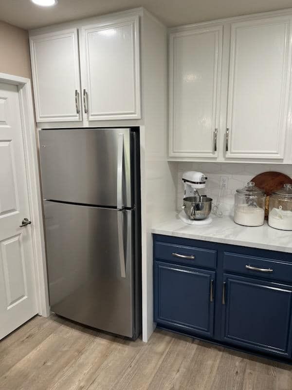 Kitchen with white upper cabinets, blue lower cabinets, stainless steel refrigerator, and wooden floors.