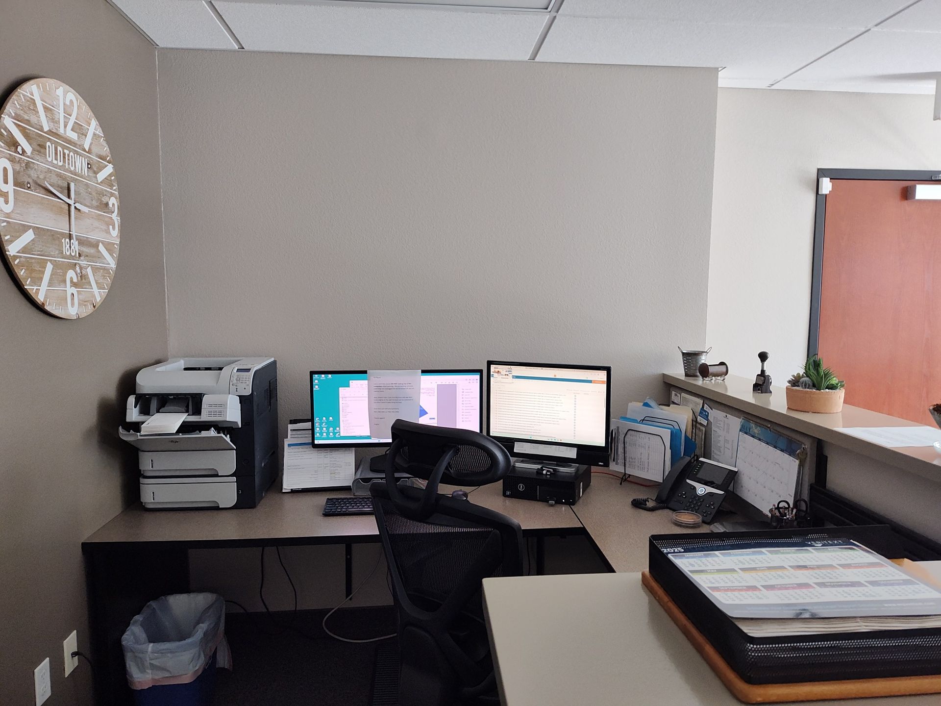 Office desk with multiple computer monitors and a printer, with a clock on the wall.