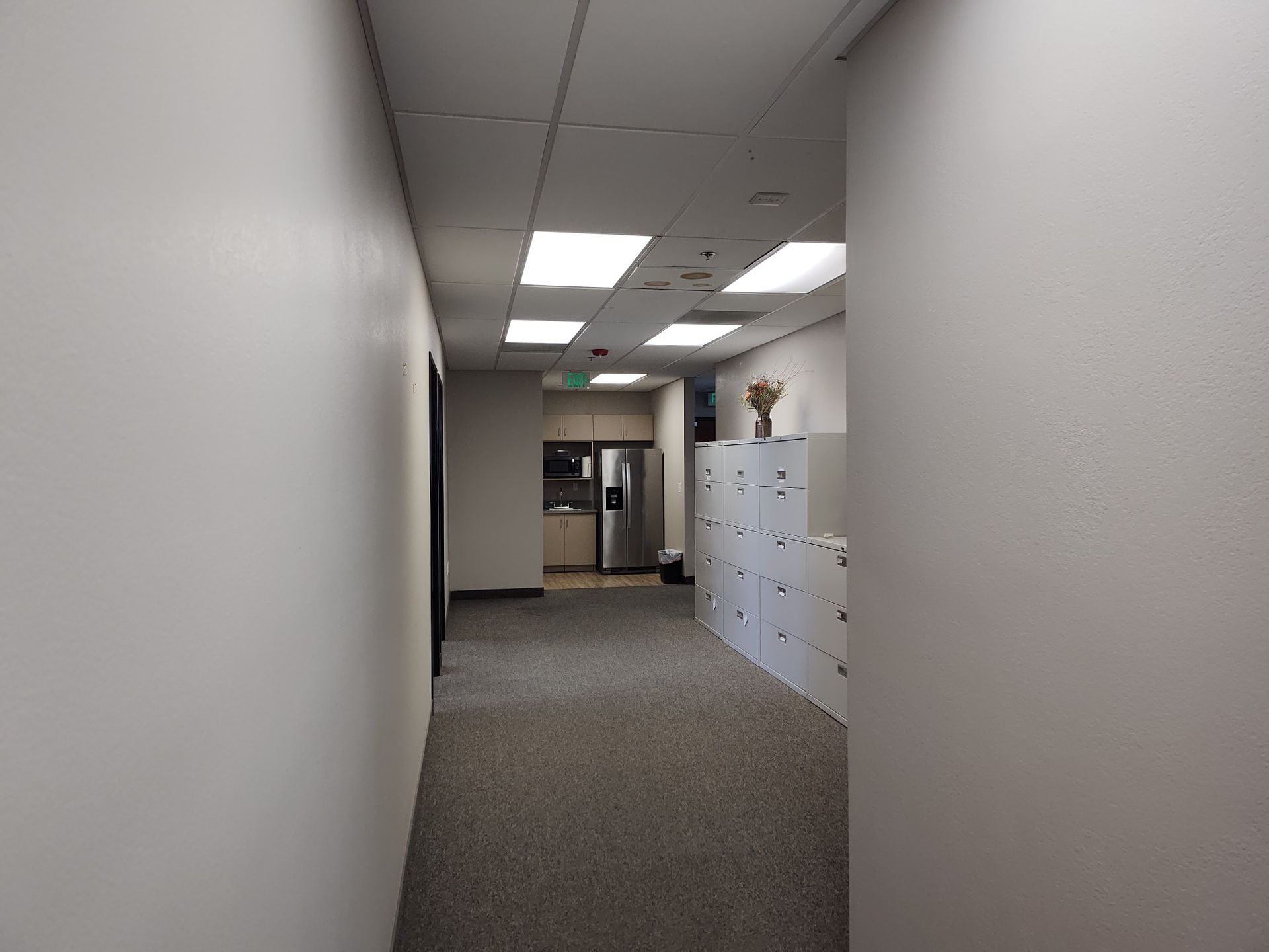 A hallway with white walls, gray carpet, and overhead lights. There are lockers on the right and a door ahead.