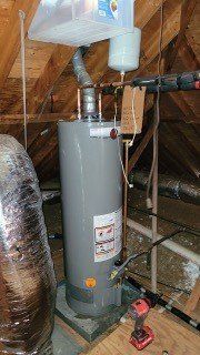 Tall gray water heater in an attic with ductwork and a red tool on the floor.