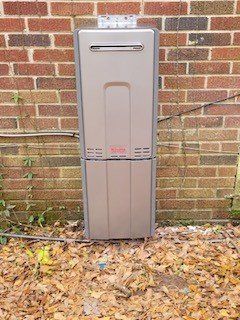 Tall gray outdoor utility cabinet mounted against a brick wall, with fallen leaves at its base.