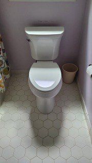 A white toilet in a small bathroom with light gray hexagon tile flooring and a trash bin beside it.