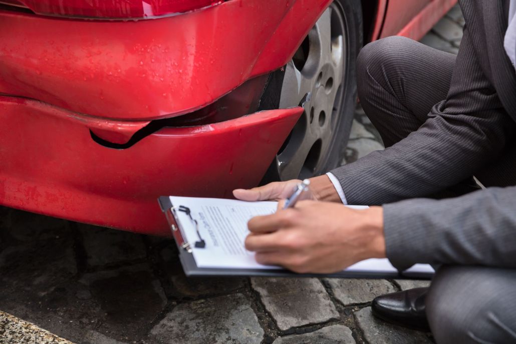 Person writes on a clipboard while inspecting a damaged red car bumper.