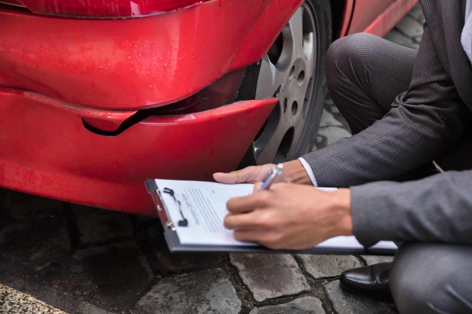 Person writes on a clipboard while inspecting a damaged red car bumper.