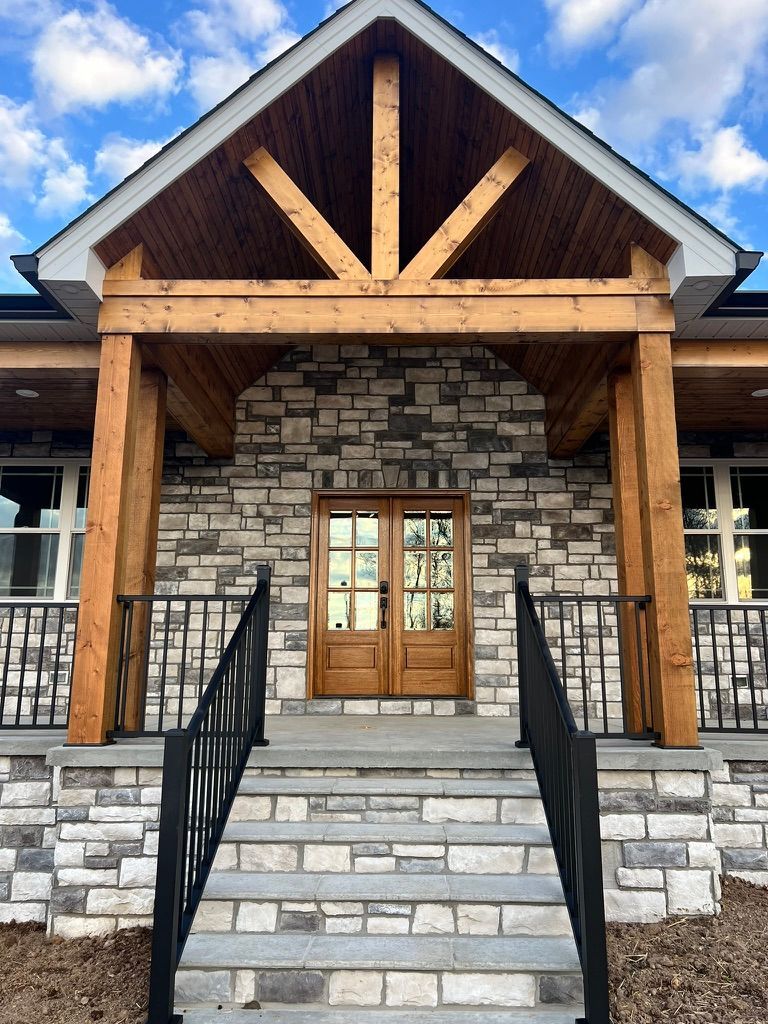Stone-faced home entrance with wooden beams, double doors, and steps leading to a porch with black railings.