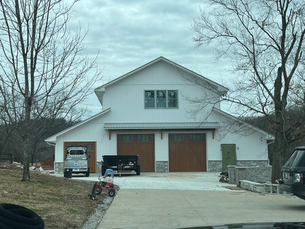 White barn with two garage doors and stone accents, parked vehicle, cloudy sky.