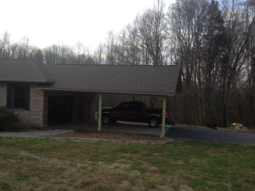 A brown pickup truck parked under a carport attached to a brick house, surrounded by trees.