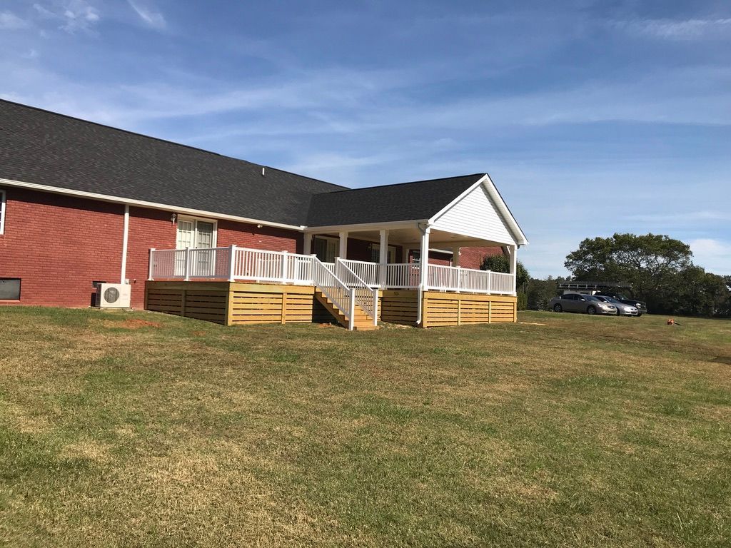 Brick house with a wooden deck and white railings on a grassy hill, with a car parked in the distance.