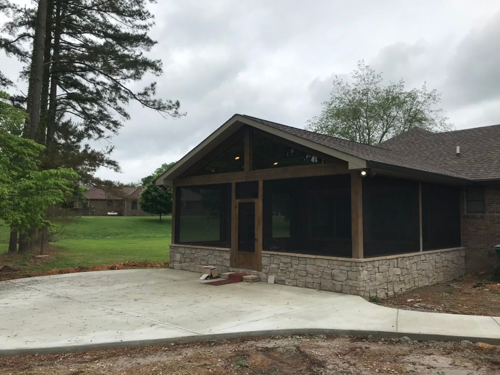 Screened porch with stone base and concrete patio, next to a brick house, under a cloudy sky.