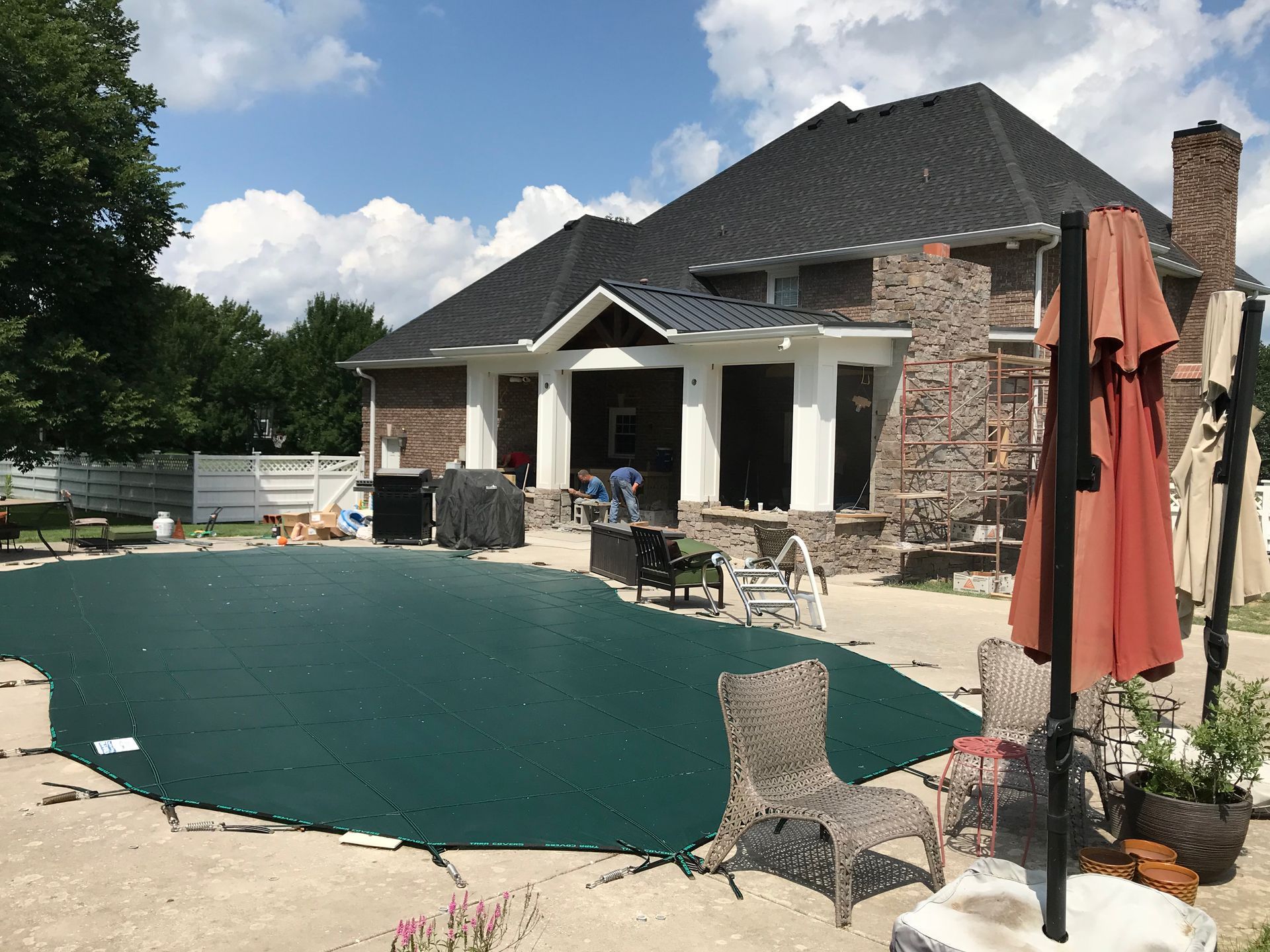 Backyard with a pool covered in green tarp. Brick house, patio furniture, and a closed umbrella.