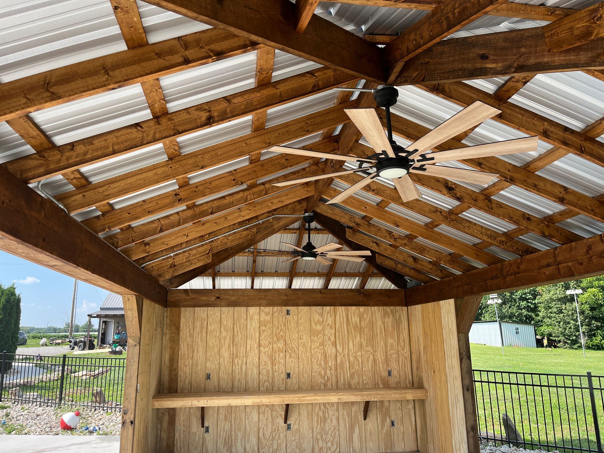 Wooden gazebo with two ceiling fans, metal roof, and plywood walls. Green grass visible outside.