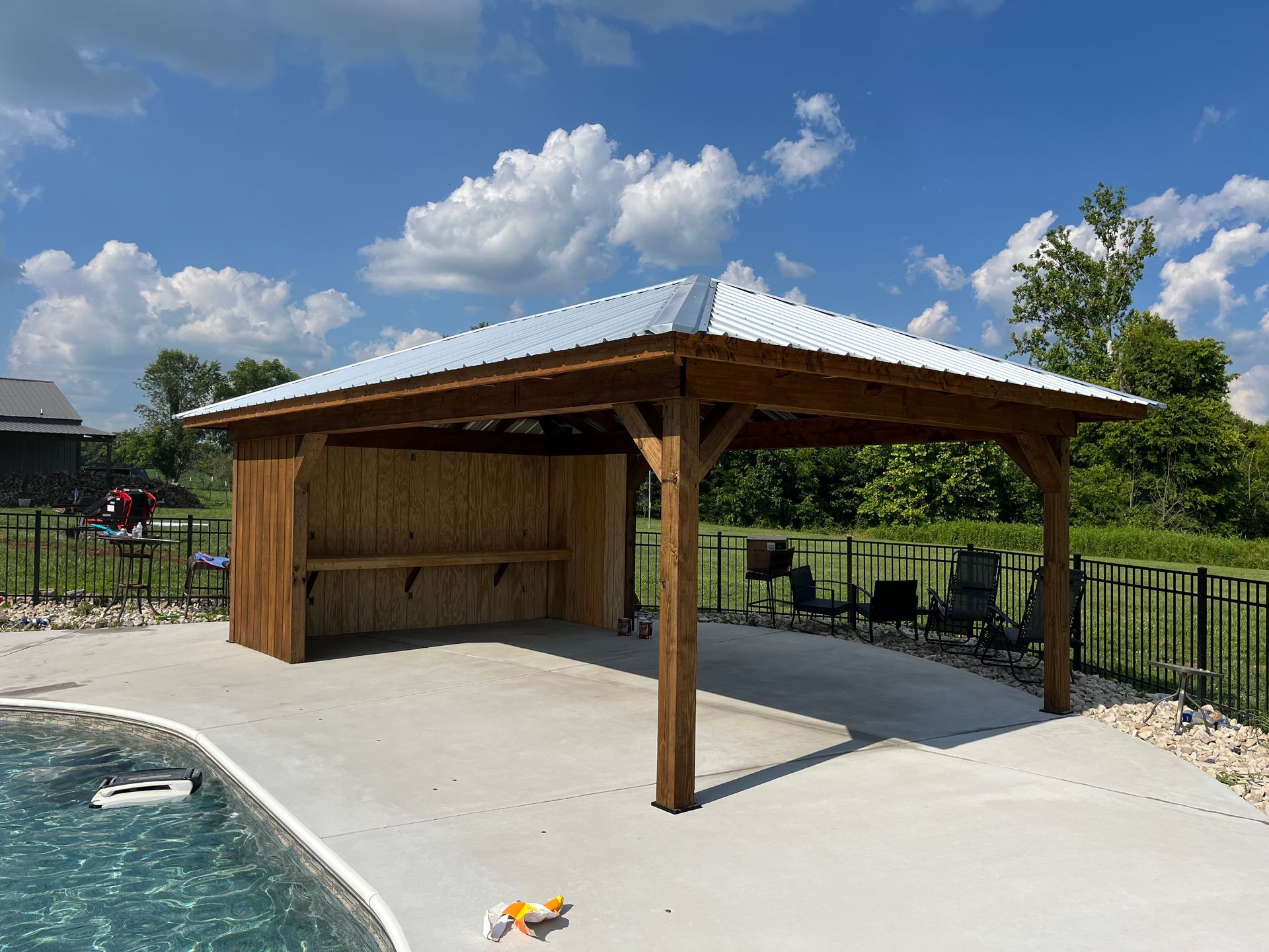 A wooden gazebo with a metal roof next to a pool, on a sunny day.