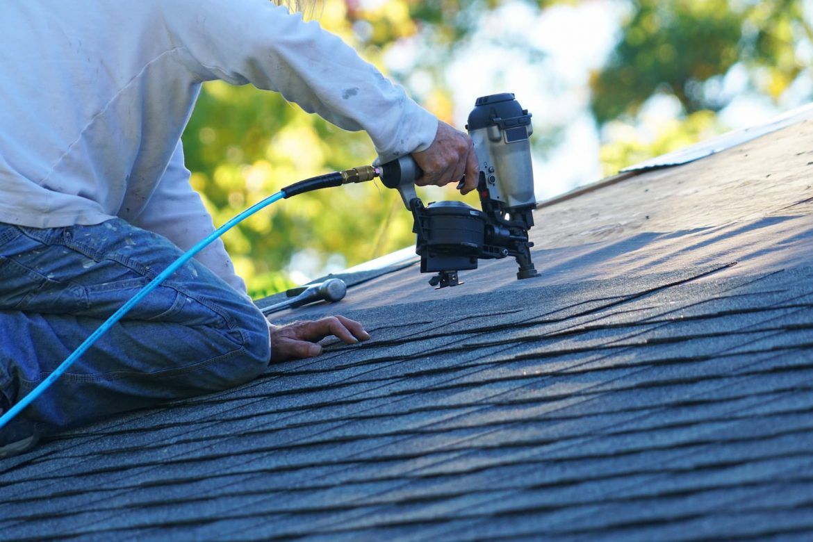 Person using a pneumatic nail gun on a dark shingle roof, blue air hose visible.