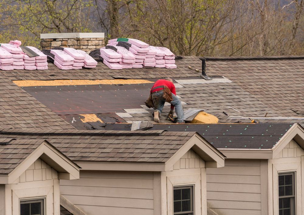 Roofer working on a roof, surrounded by shingles and insulation. Gray and brown roof, tan house, and trees in background.