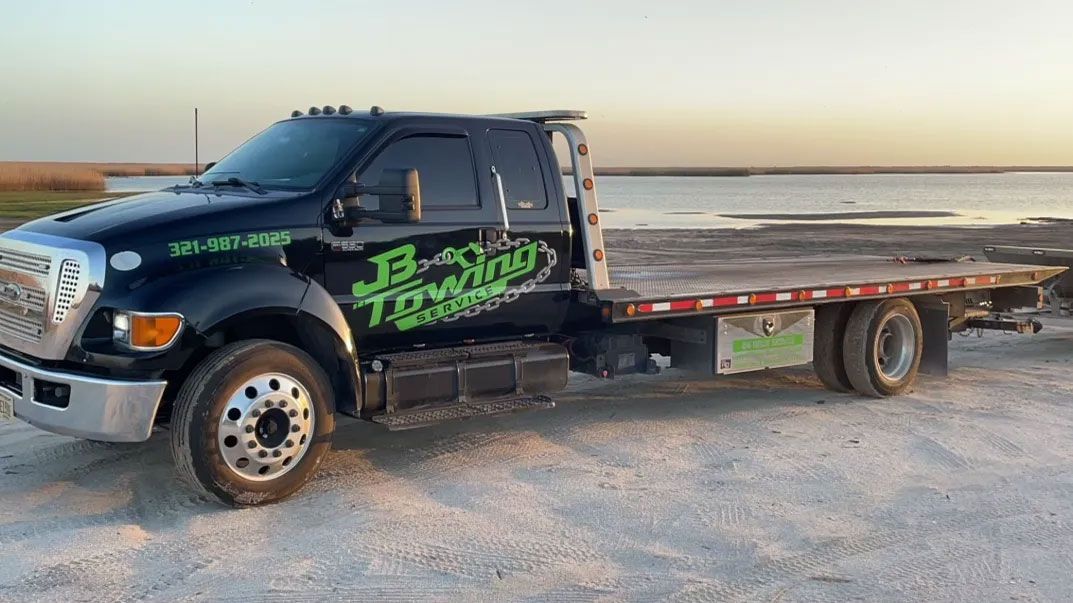 Black flatbed tow truck on a sandy beach at sunset, facing left