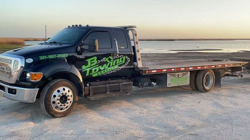 Black tow truck parked on a beach at sunset, with a flatbed and green logo on the door