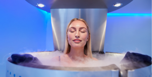 Woman in a cryotherapy chamber, eyes closed, with vapor around her shoulders. Blue lighting.
