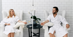 A woman and a man in white robes receive IV infusions while sitting in armchairs. They are smiling and looking at each other.