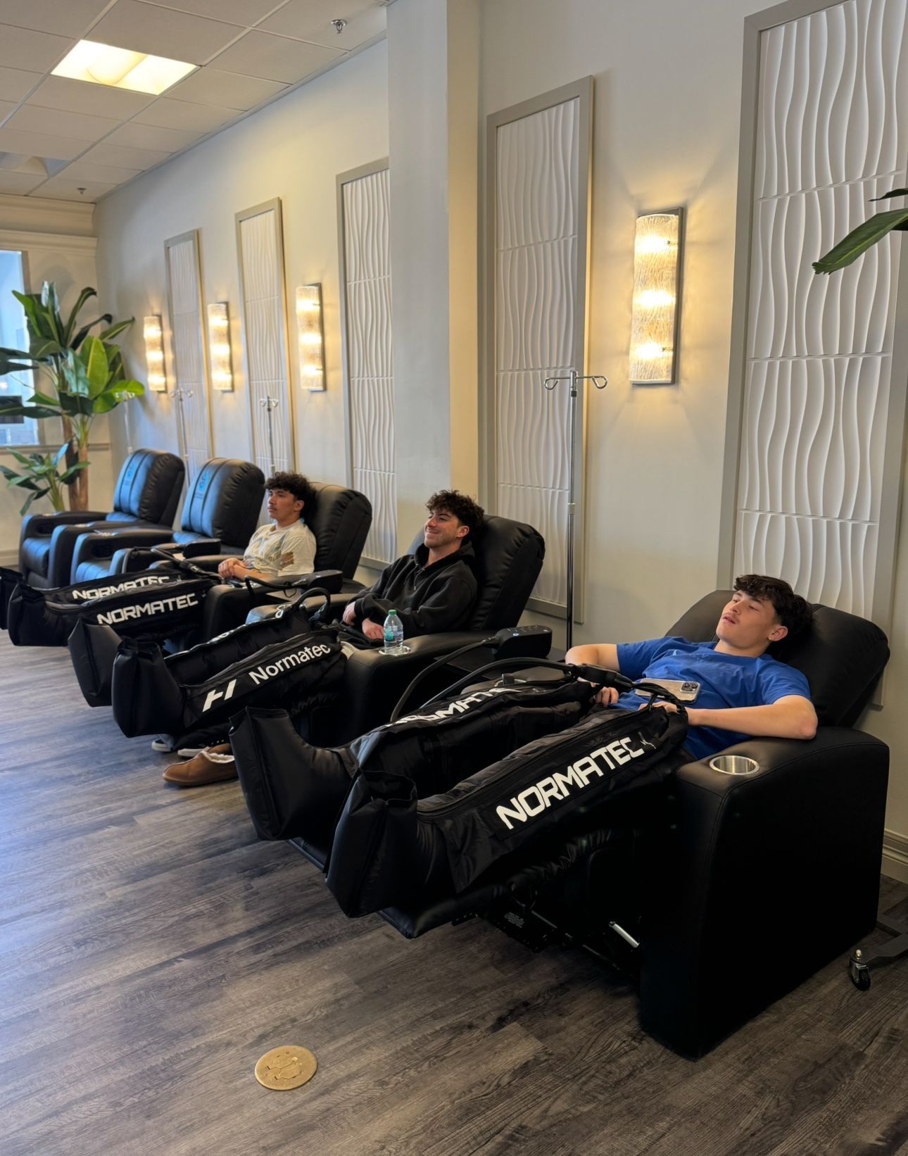 Three people relaxing in massage chairs with Normatec leg sleeves in a recovery lounge.