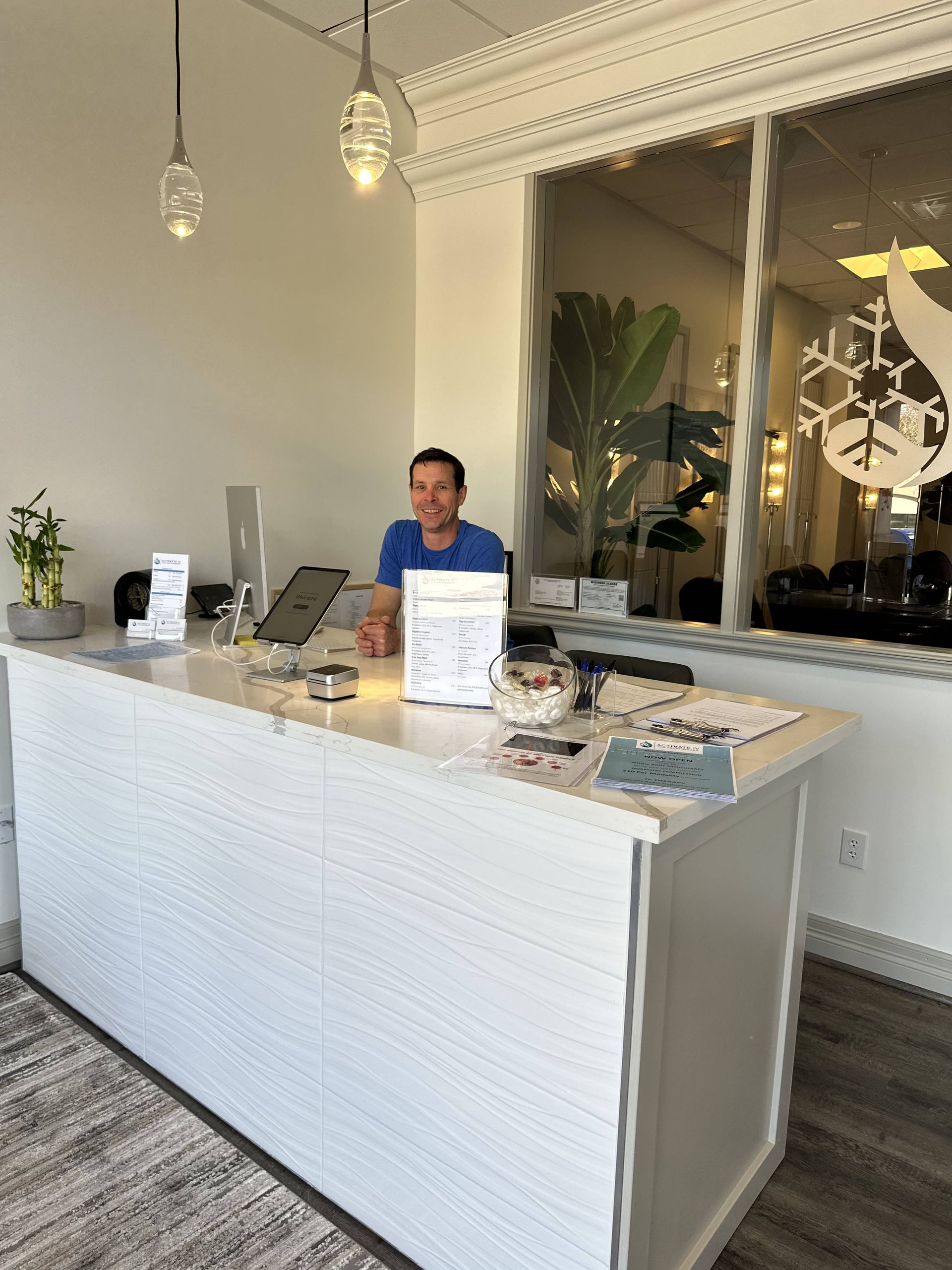 Person smiling behind a white reception desk in a brightly lit office. Decorative lights and a large window are visible.