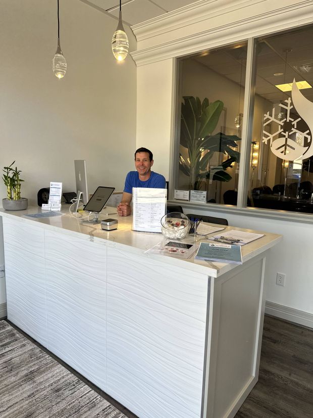 Person smiling behind a white reception desk in a brightly lit office. Decorative lights and a large window are visible.
