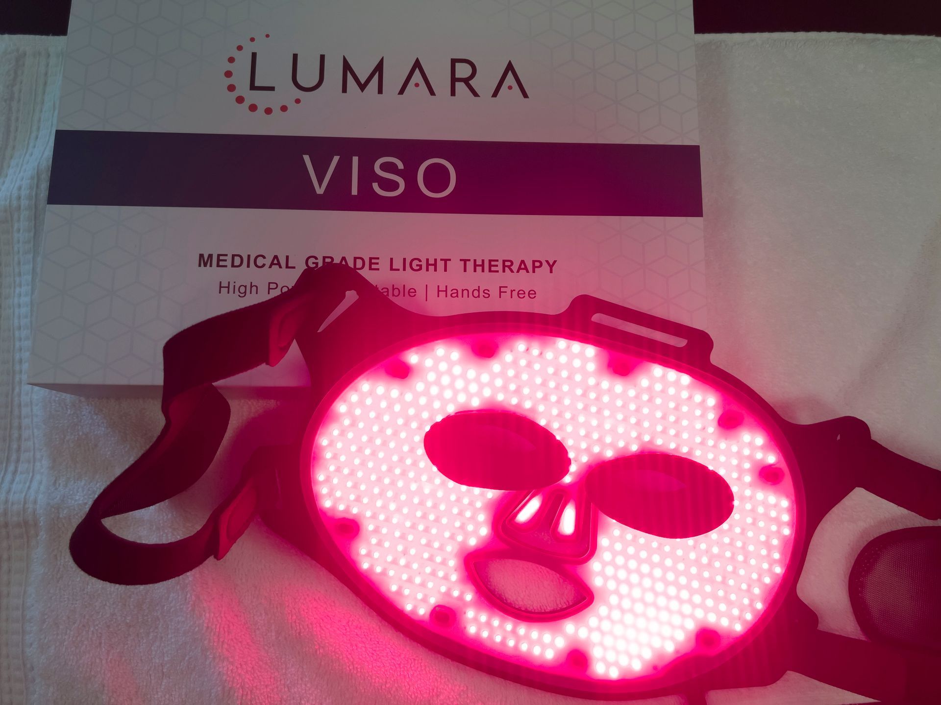 A Lumara Viso LED light therapy mask illuminated in red light, with its box in the background.