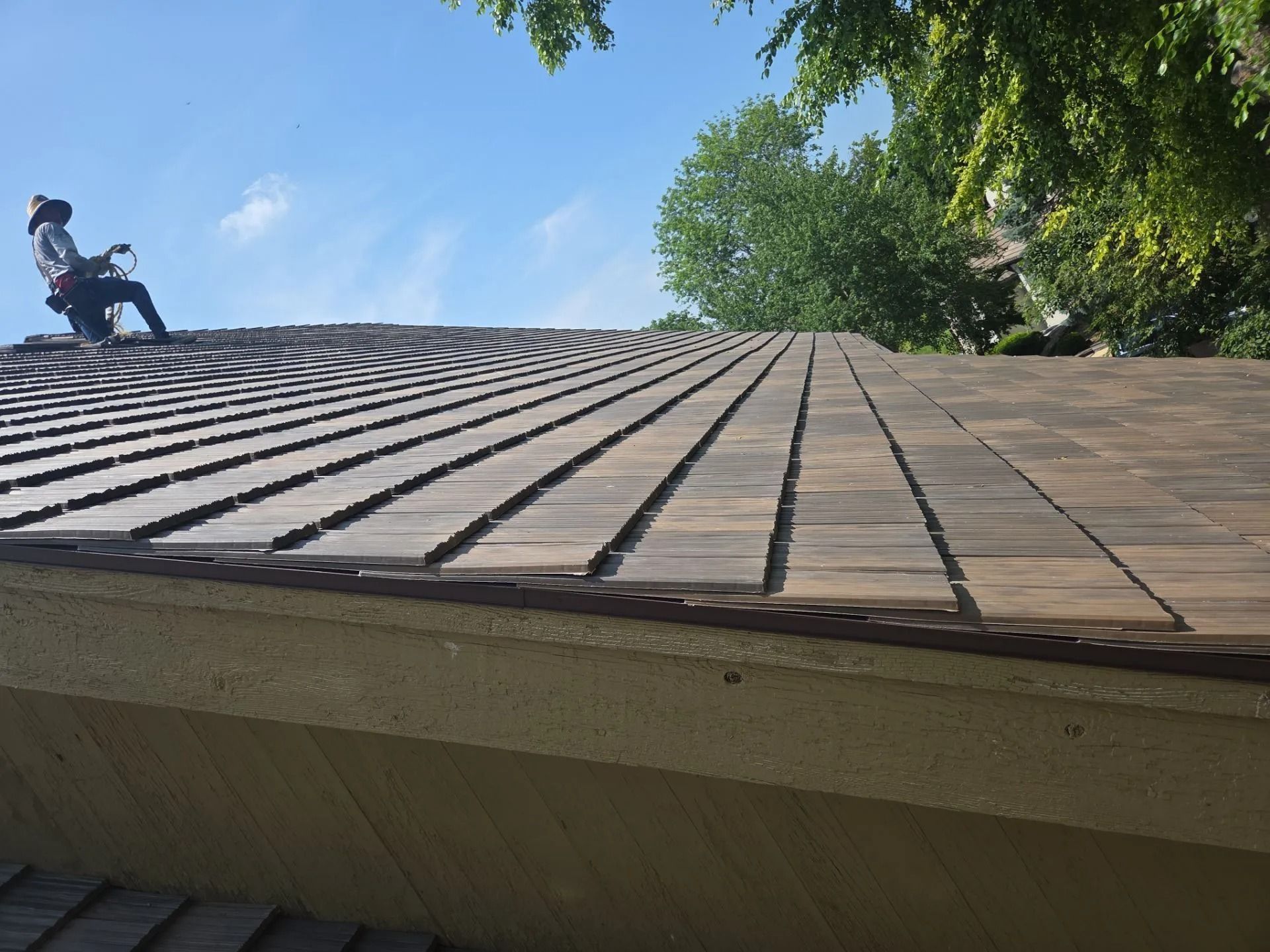 Roofer on a shingled roof, blue sky background. The roof is partially repaired.