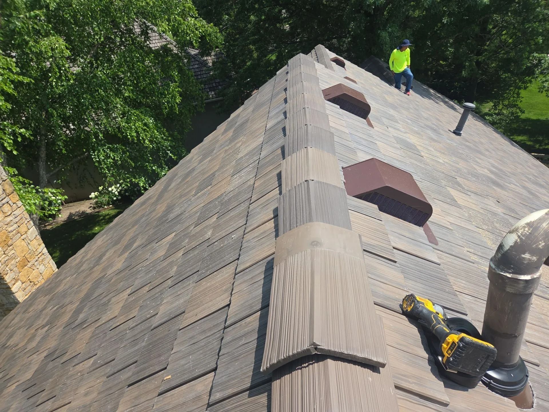 Roofer on a shingle roof with vents; electric drill on the roof. Sunny day, trees in background.