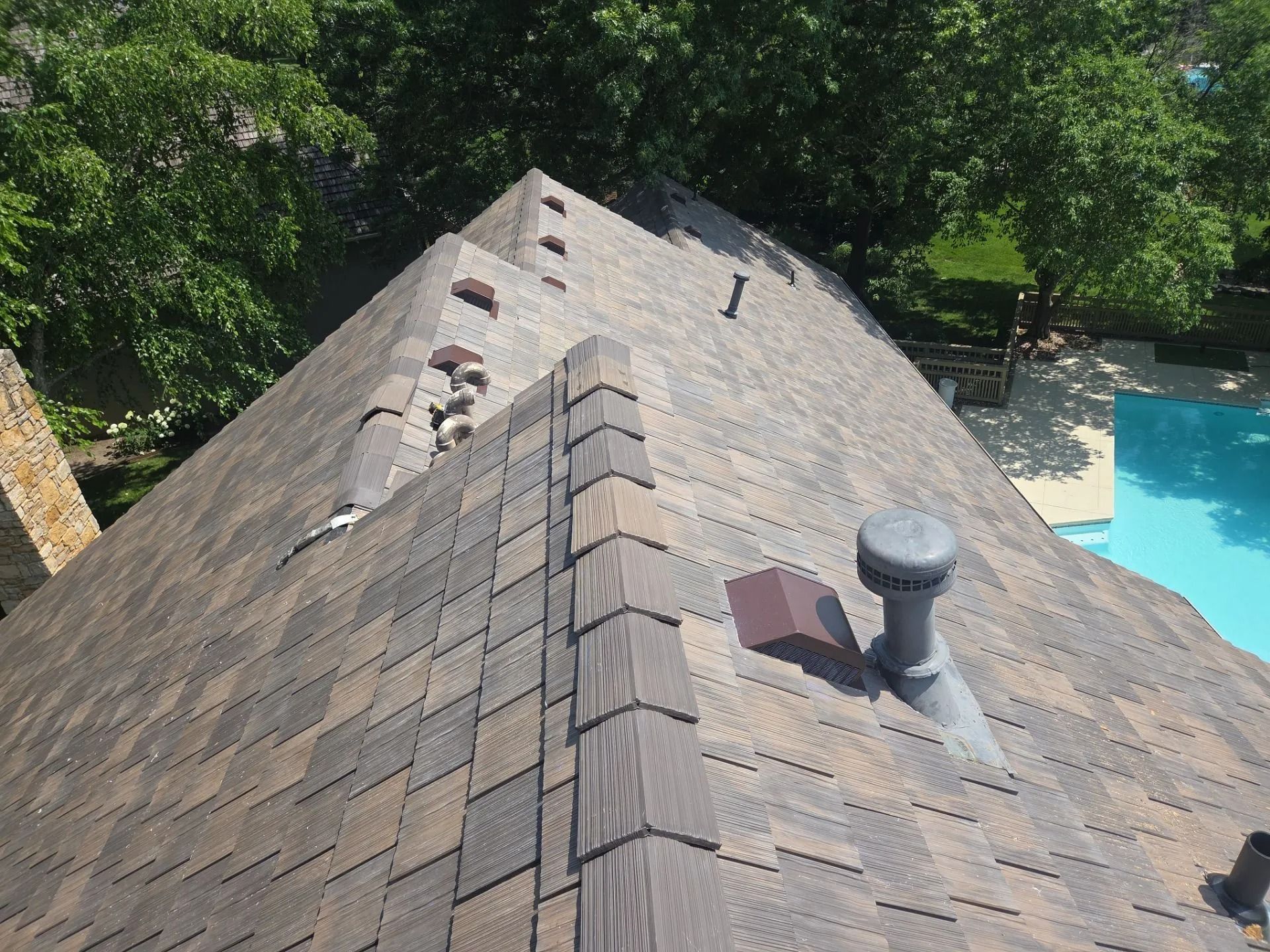 Overhead view of a house roof with brown shingles, chimney, and a glimpse of a pool.