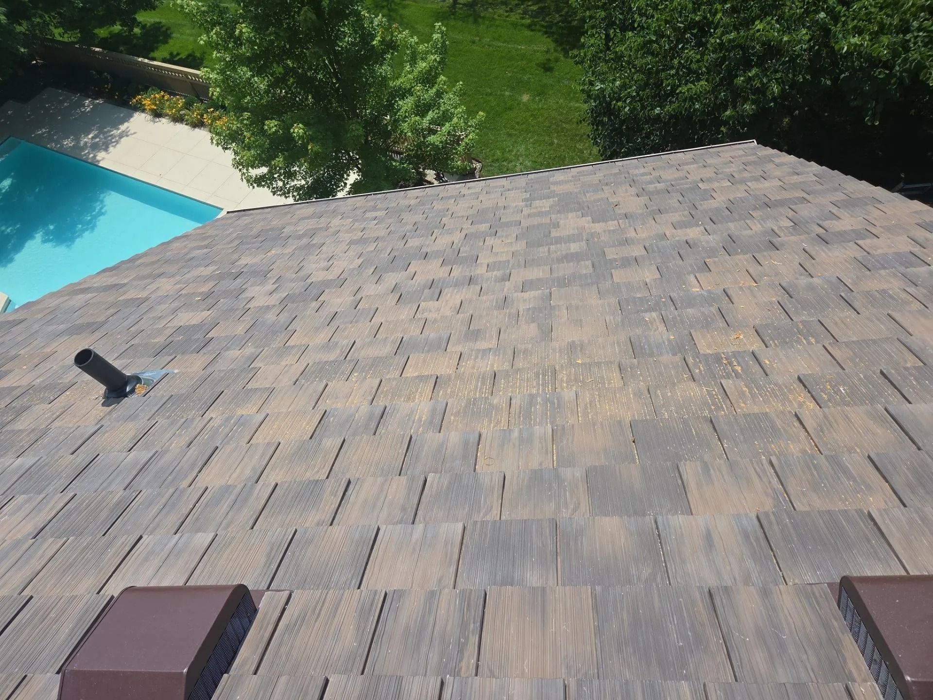 View of a brown and gray shingled roof with a swimming pool and trees in the background.