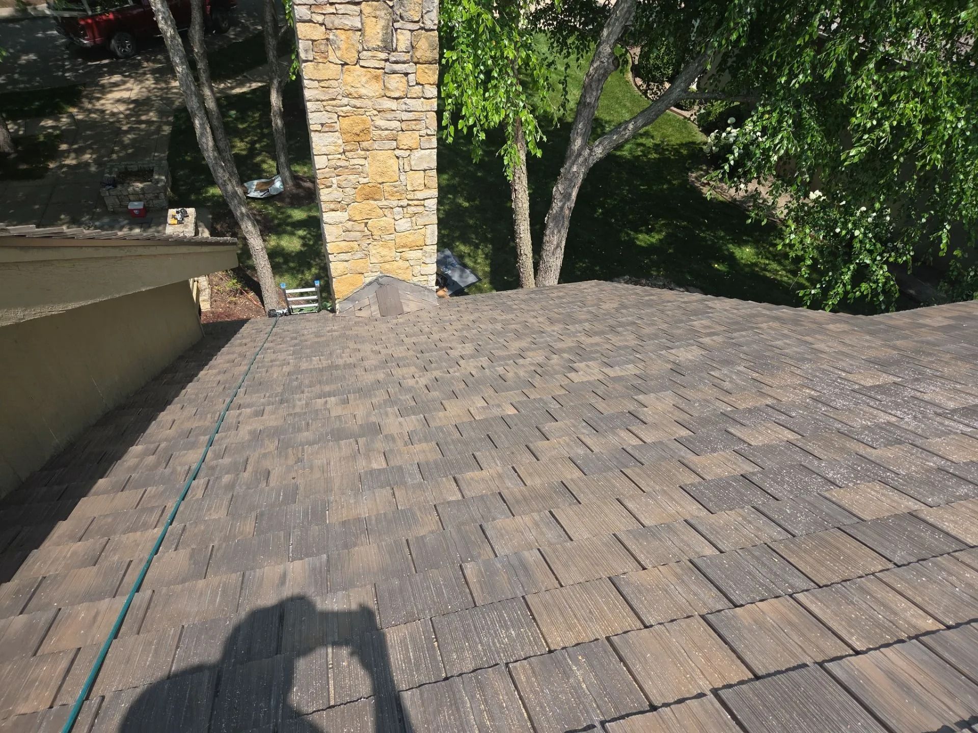 View from a roof, showing brown asphalt shingles, chimney, and trees in background.