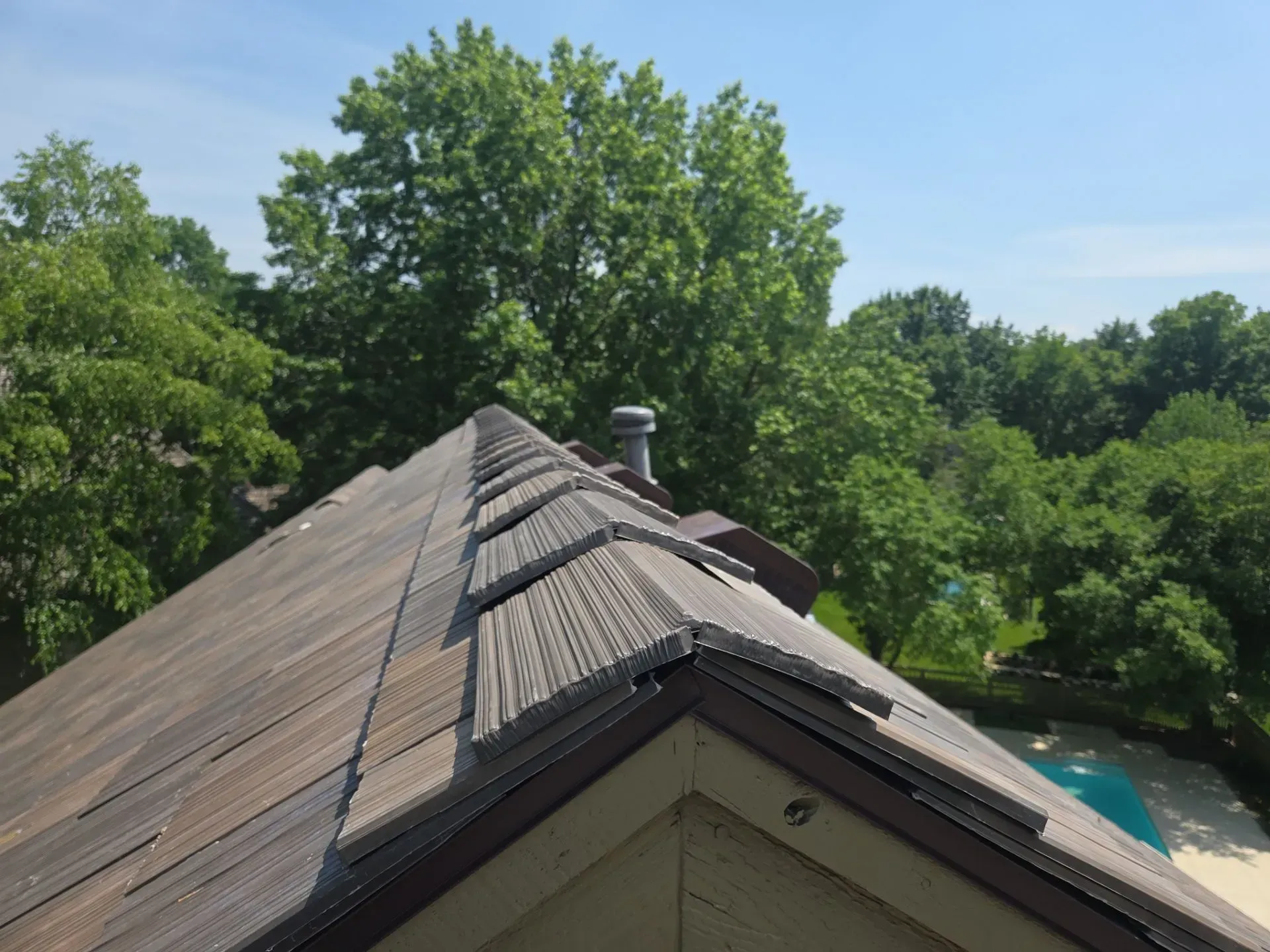 Damaged asphalt shingle roof on a house, with missing and curling shingles, trees and pool visible.