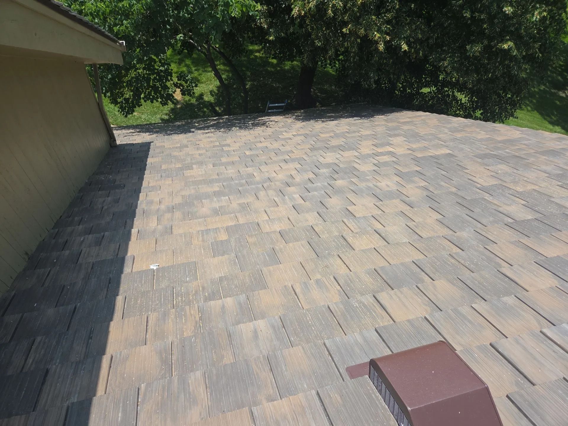 Close-up of a brown and gray shingled roof with a vent in front of a tree line.