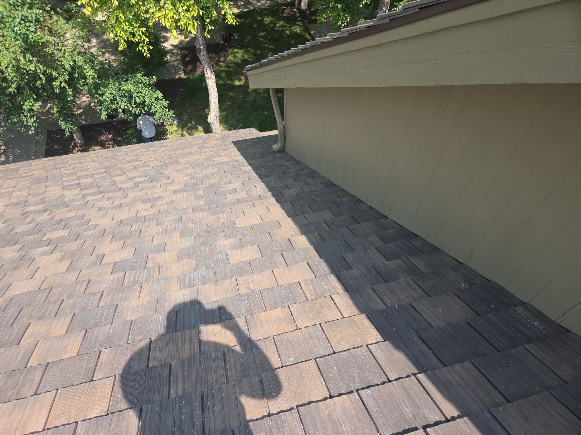 Shadow of a person taking photo on a shingled roof next to a light-colored wall and trees.