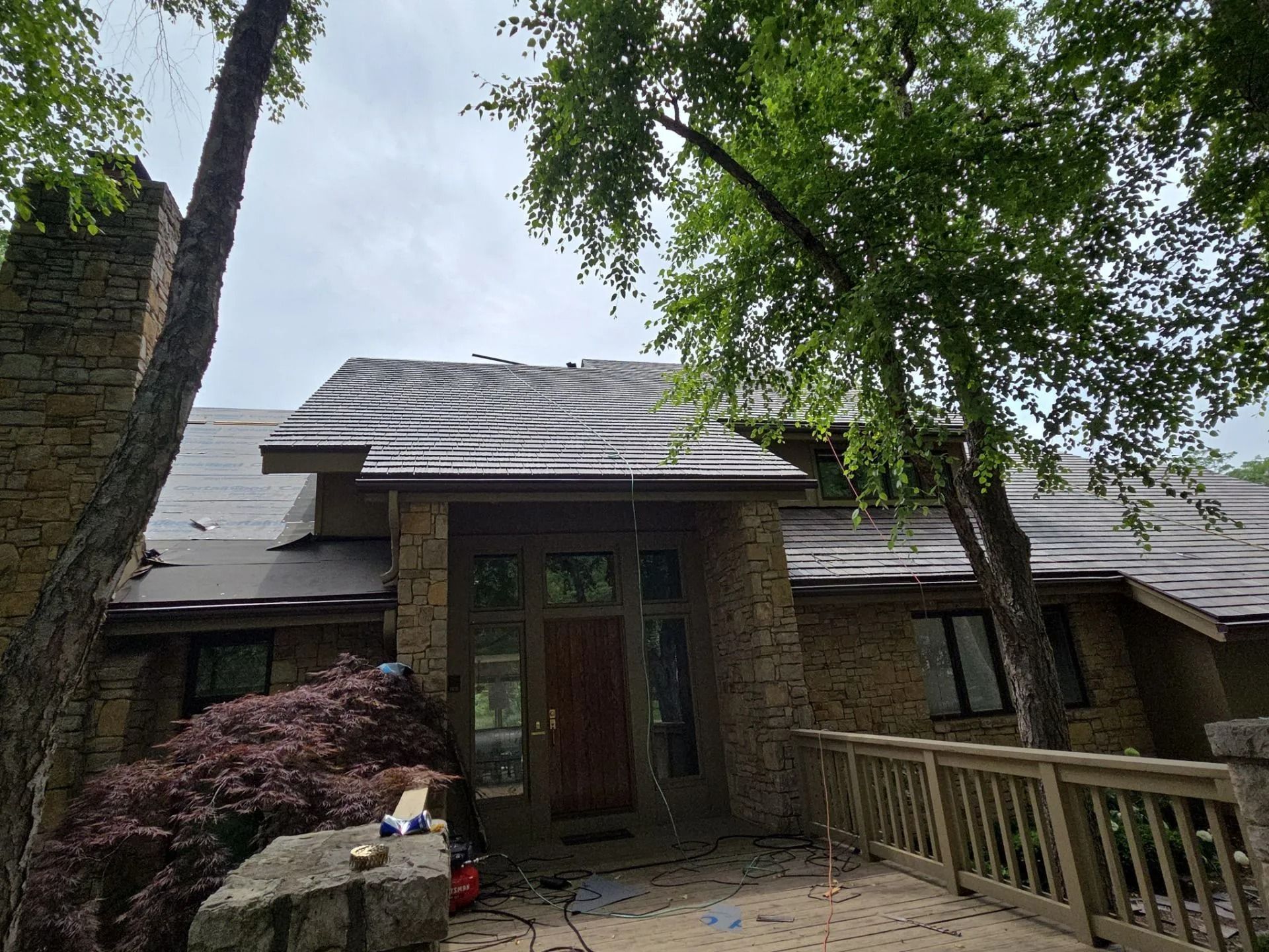 House with damaged roof and exposed underlayment; stone facade, trees, cloudy sky.