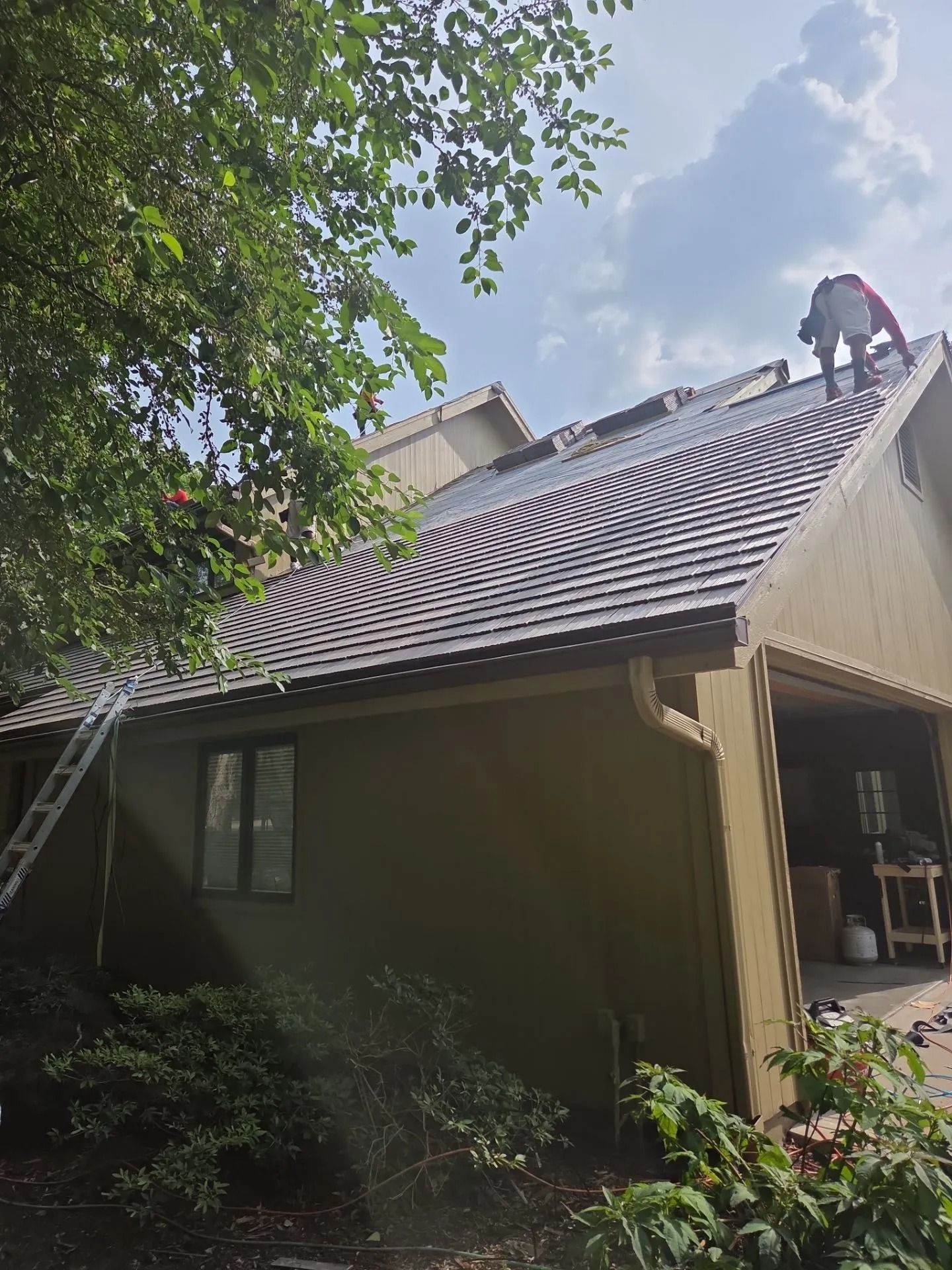 Person on a roof installing tiles on a house. Green bushes and a ladder are visible. Sunny day.