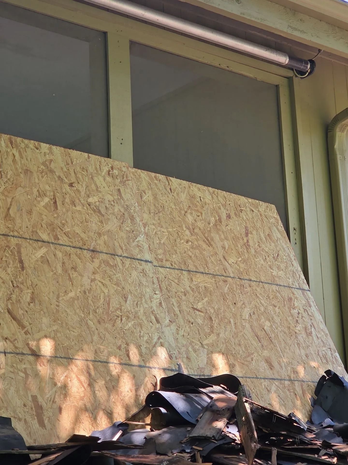 Plywood board covers a window, debris below. A light fixture is mounted above.