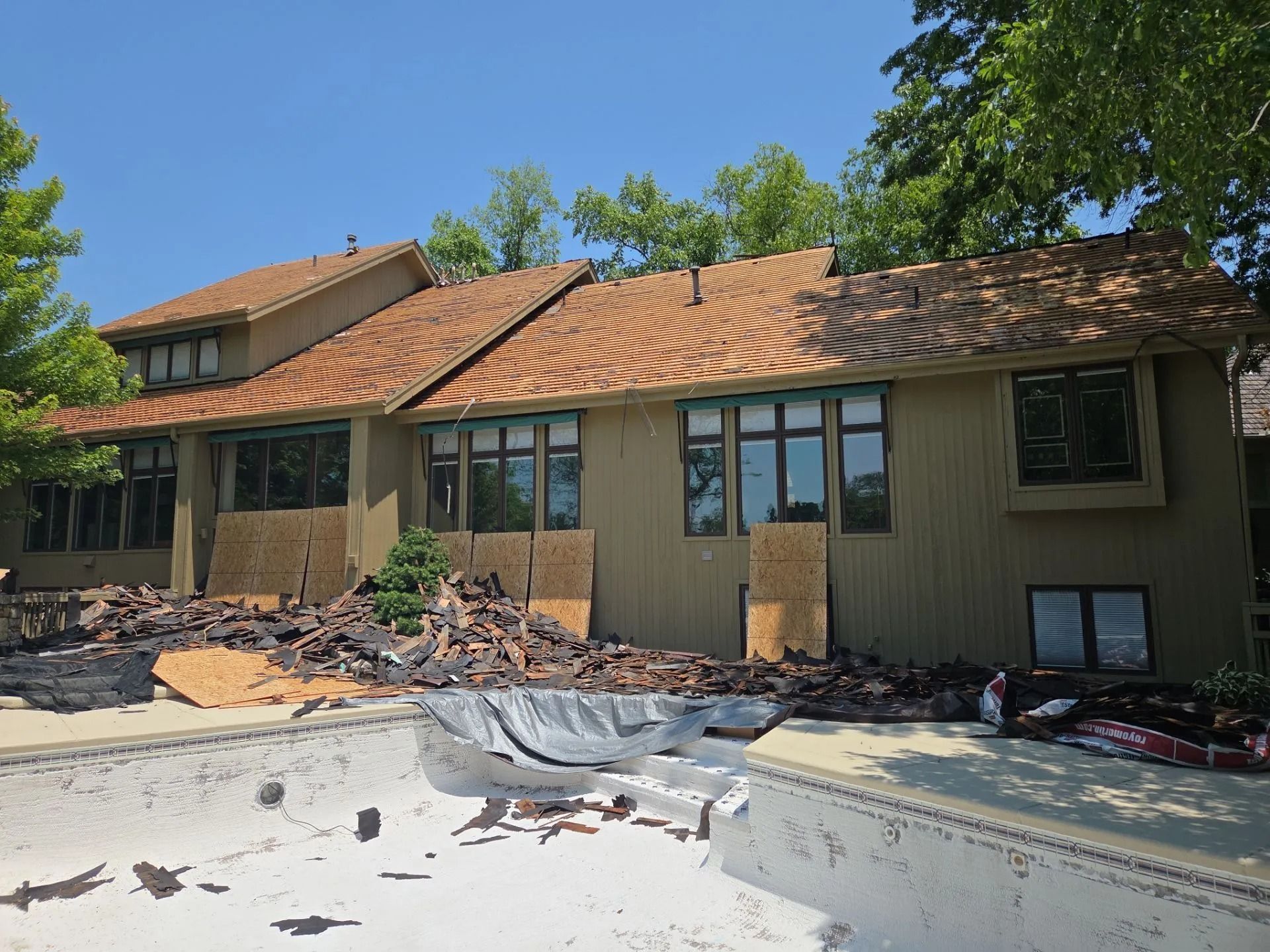 House with roof damage, debris in pool. Roof shingles removed, plywood covers windows, sunny day.