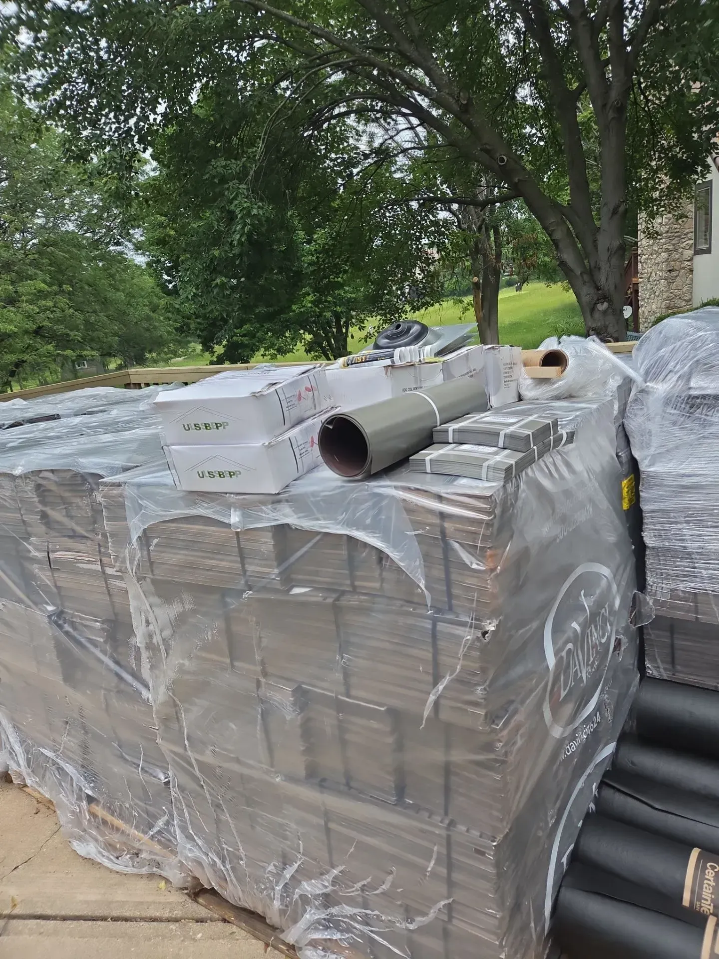 Stacked building materials wrapped in plastic, outdoors. Includes gray siding, pipes, and boxes.
