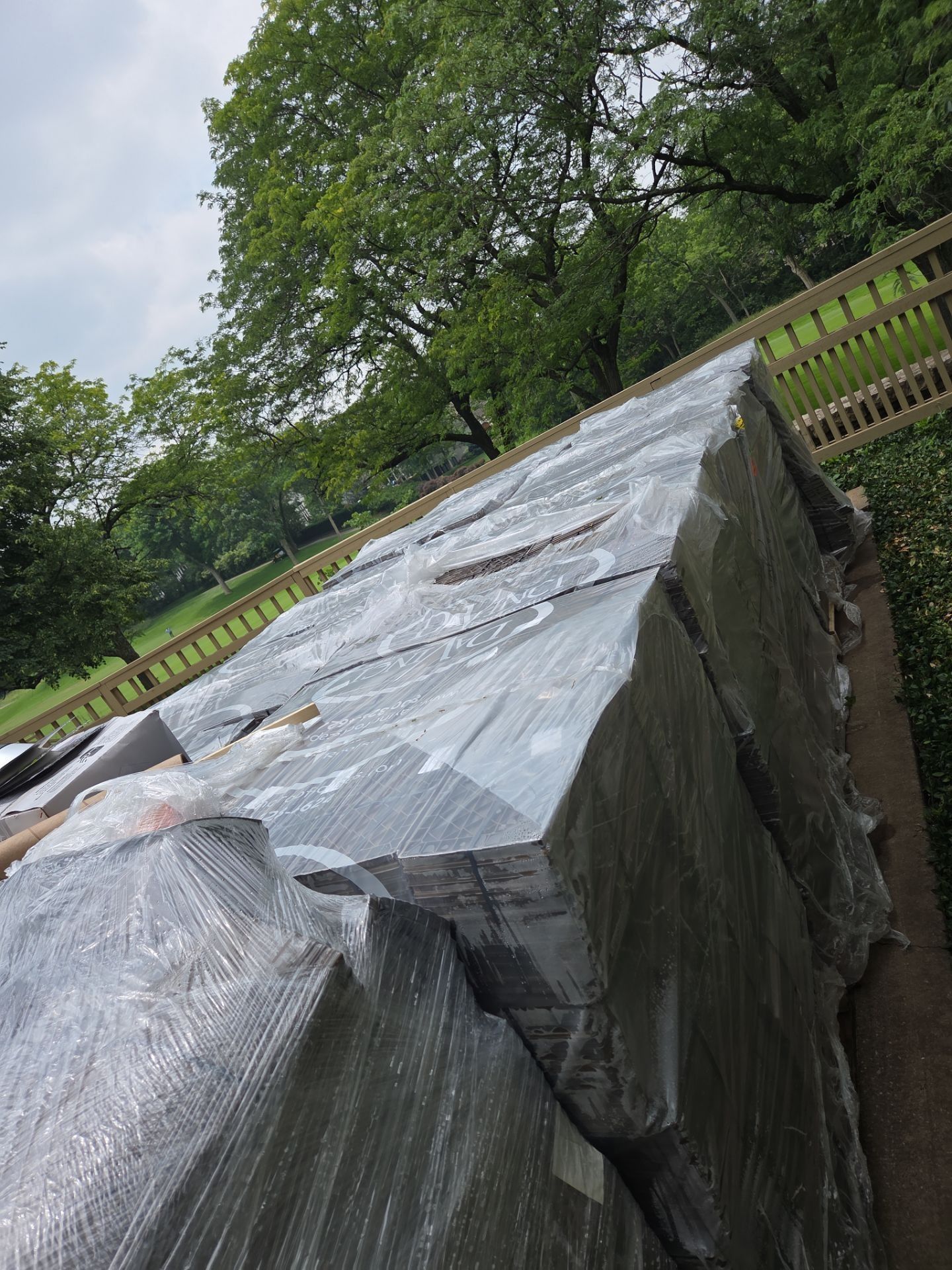 Large, wrapped pallets of dark-colored material in an outdoor setting, near a wooden fence and trees.