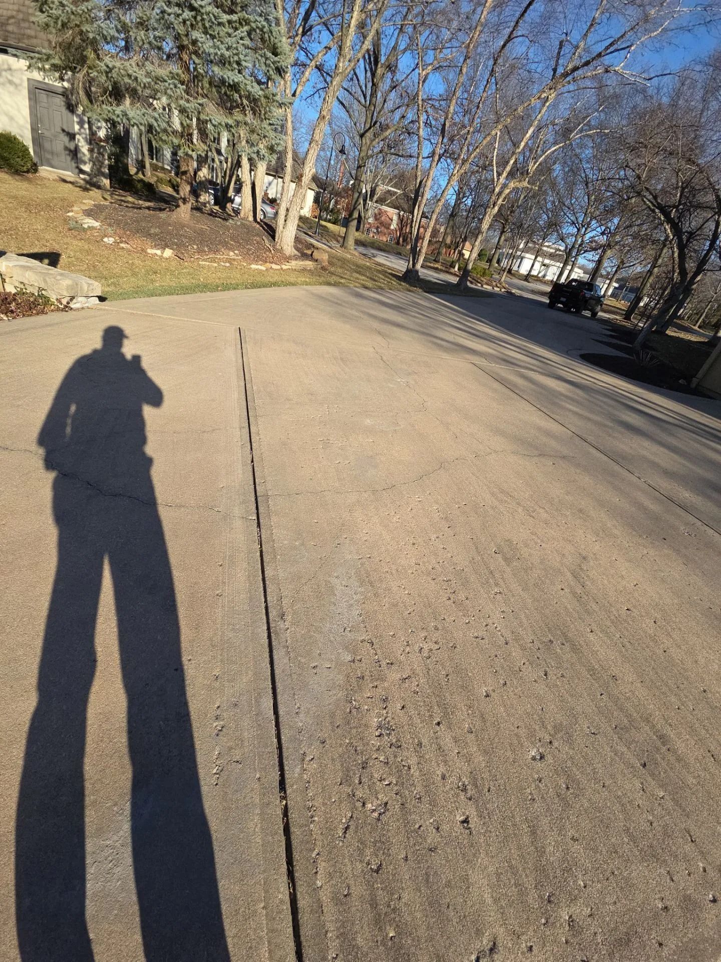Shadow of a person on a cracked concrete driveway, trees and houses in the background.