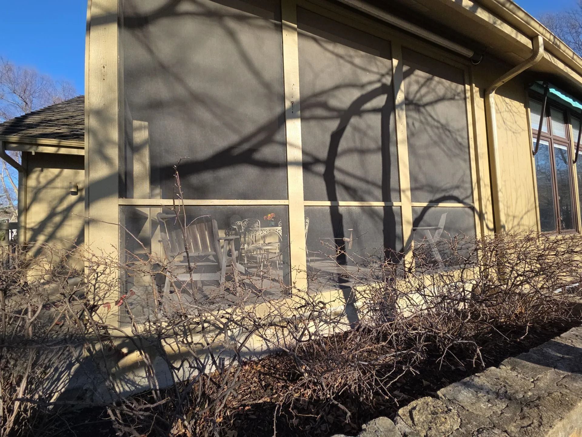 Screened porch with bare branches casting shadows. Beige walls, brown roof, and stone wall.