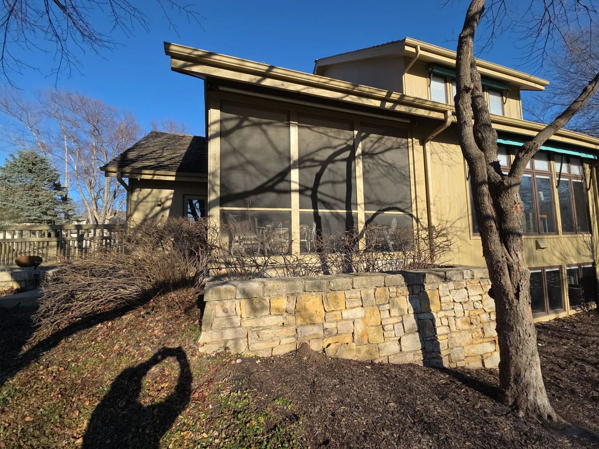 Tan house with screened porch, stone retaining wall, leafless trees against a blue sky.