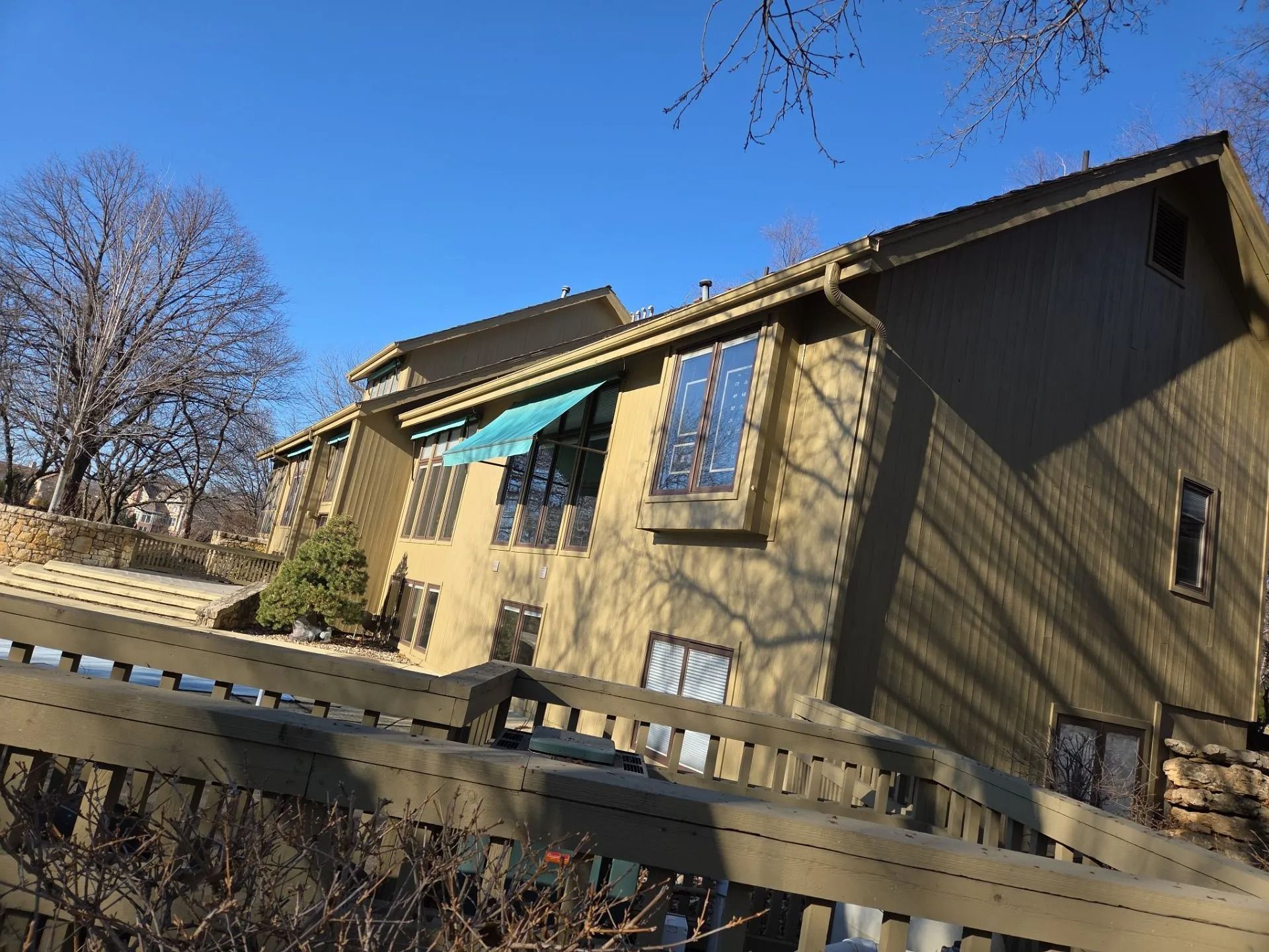 Two-story brown house with a blue awning on a sunny day. A wooden fence is in the foreground.