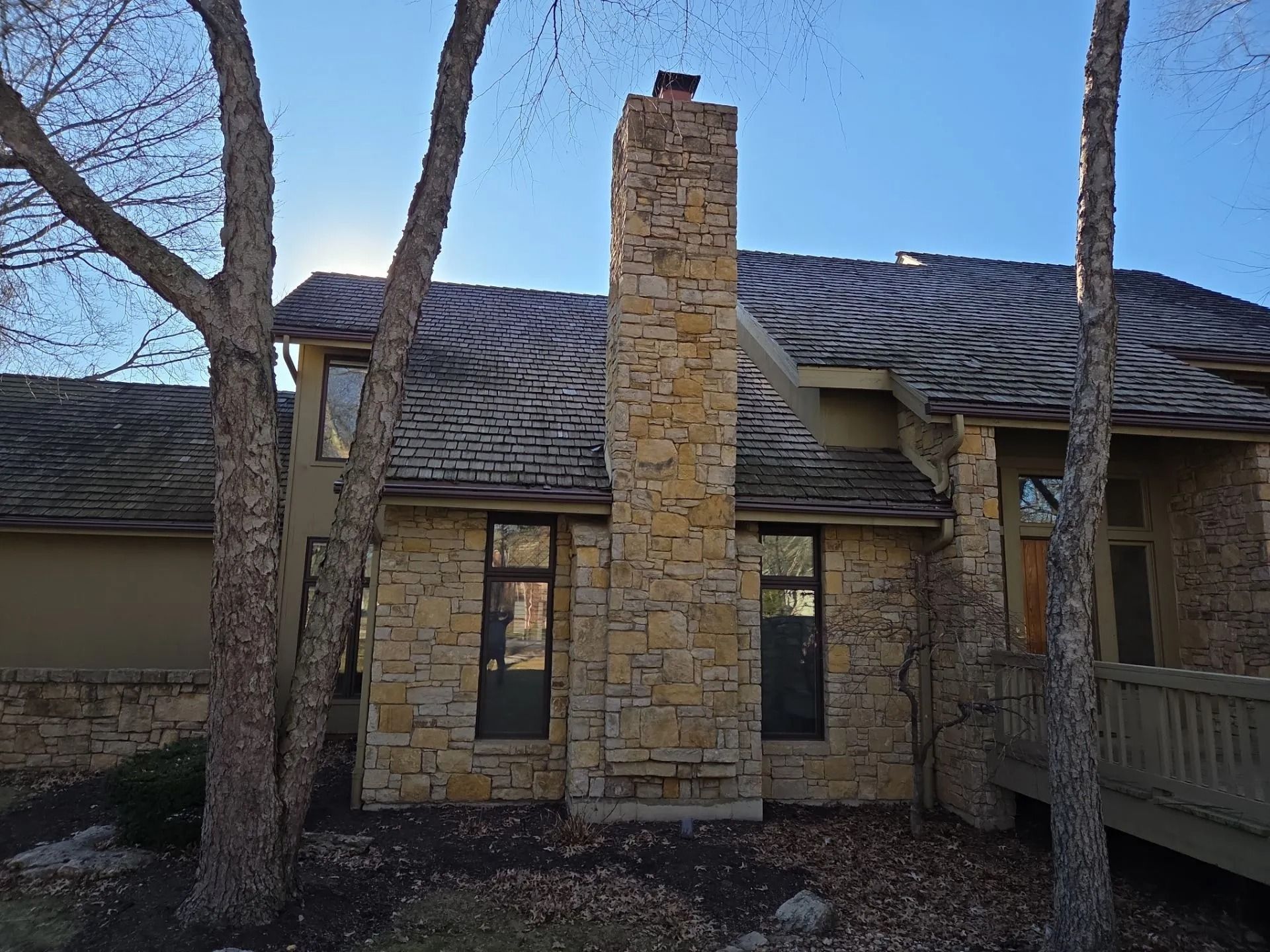 Stone chimney and house exterior with windows, trees, and wooden deck under a blue sky.