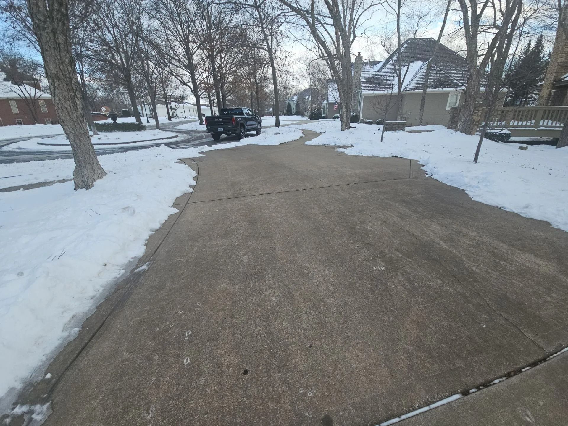 Snowy driveway with a black truck, trees, and houses in the background.